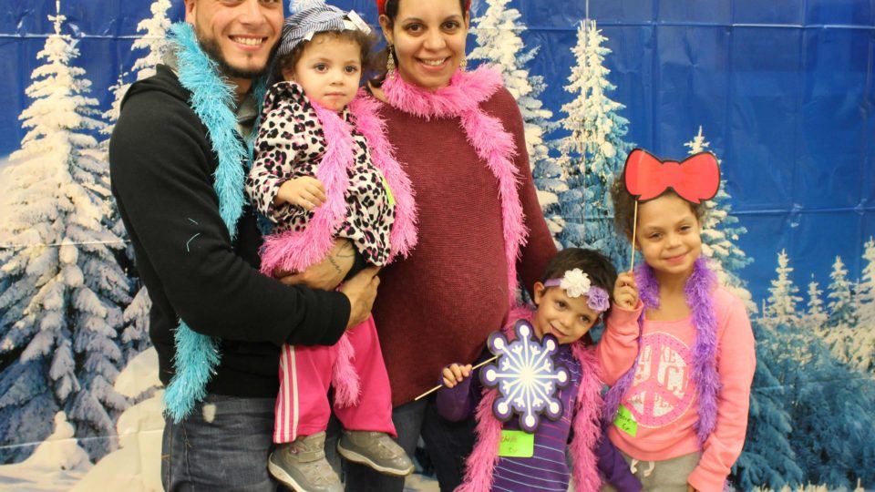 Pediatric patient with siblings and caregivers pose in front of winter themed photo booth backdrop during holiday Party Time program